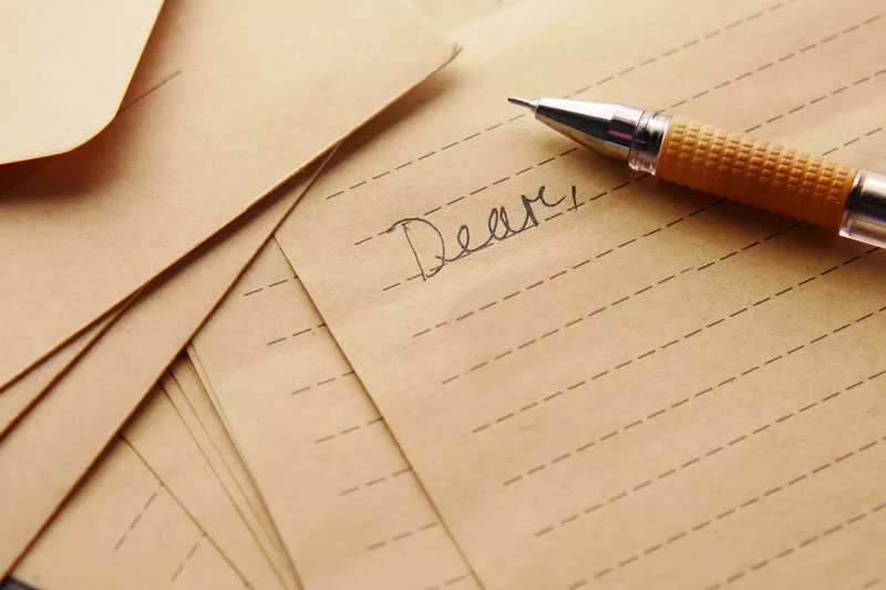 Kraft-brown letter paper ruled with dashed lines, the word Dear written in ink, a pen across the corner