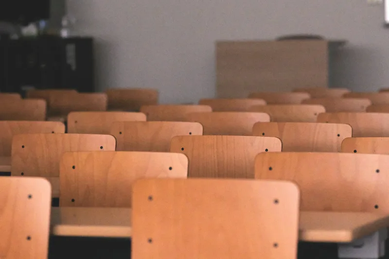 Row upon row of empty pale beech chair backs in a bright teaching room, the far rows in soft focus